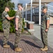 Three U.S. Marines in MARPAT woodland camouflage uniforms are gathered on a brick plaza in an outdoor military installation setting. One Marine on the left appears to be pinning an insignia or award onto the center Marine's uniform collar or chest area, suggesting a promotion or award ceremony. A third Marine stands at attention facing them, likely serving as a witness to the formal proceeding.