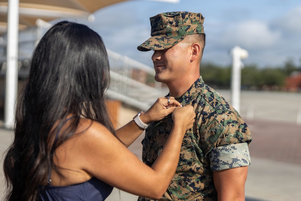 A woman is pinning an insignia or device onto the collar/chest area of a male U.S. Marine in MARPAT digital camouflage uniform. The setting appears to be an outdoor military installation or parade ground. The scene is consistent with a military promotion or pinning ceremony.