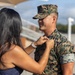 A woman is pinning an insignia or device onto the collar/chest area of a male U.S. Marine in MARPAT digital camouflage uniform. The setting appears to be an outdoor military installation or parade ground. The scene is consistent with a military promotion or pinning ceremony.