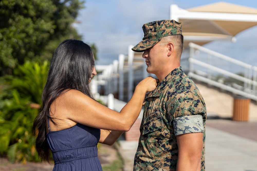 A woman in a navy blue dress is pinning something onto the collar or chest area of a U.S. Marine in MARPAT digital camouflage uniform. The setting appears to be an outdoor military base or installation, likely during a promotion or award ceremony. The interaction suggests a personal and ceremonial moment, possibly a rank promotion pinning.