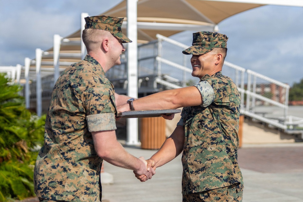 Two U.S. Marines in MARPAT woodland camouflage uniforms are engaged in a formal handshake ceremony outdoors. One Marine is presenting a plaque or award to the other, who is smiling broadly. The setting appears to be a military installation with bleachers and shade structures visible in the background.