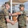 Chief Warrant Officer 2 Joshua J. Krantz’s Promotion Ceremony Two U.S. Marines in MARPAT woodland camouflage uniforms are engaged in a formal handshake ceremony outdoors. One Marine is presenting a plaque or award to the other, who is smiling broadly. The setting appears to be a military installation with bleachers and shade structures visible in the background.