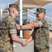 Two U.S. Marines in MARPAT woodland camouflage uniforms are engaged in a formal handshake ceremony outdoors. One Marine is presenting a plaque or award to the other, who is smiling broadly. The setting appears to be a military installation with bleachers and shade structures visible in the background.