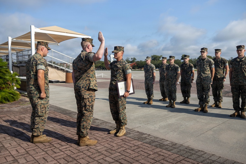 A military ceremony is taking place outdoors on a paved parade ground, likely a promotion or award ceremony. One Marine is raising his right hand in an oath while holding a framed certificate, with a senior Marine officiating in front of him. A formation of Marines stands at attention in the background as witnesses.