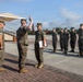 A military ceremony is taking place outdoors on a paved parade ground, likely a promotion or award ceremony. One Marine is raising his right hand in an oath while holding a framed certificate, with a senior Marine officiating in front of him. A formation of Marines stands at attention in the background as witnesses.