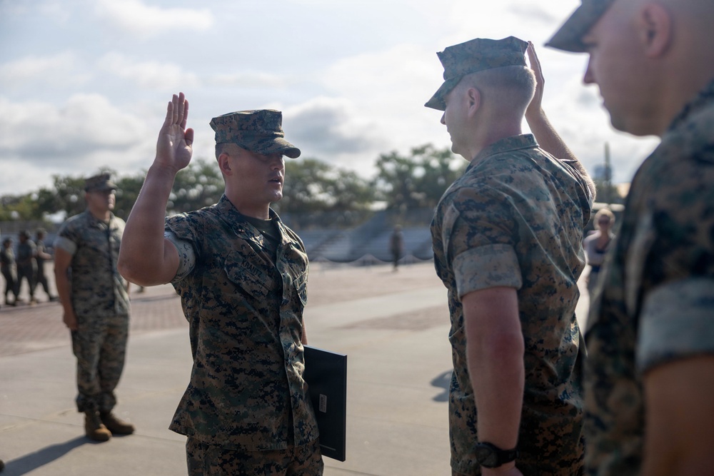 A military ceremony appears to be taking place outdoors on a paved area, likely a promotion or re-enlistment ceremony. A Marine in the center is raising his right hand while holding a folder, suggesting he is taking an oath, while another Marine renders a salute. Additional uniformed personnel are visible in the background.