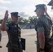 A military ceremony appears to be taking place outdoors on a paved area, likely a promotion or re-enlistment ceremony. A Marine in the center is raising his right hand while holding a folder, suggesting he is taking an oath, while another Marine renders a salute. Additional uniformed personnel are visible in the background.