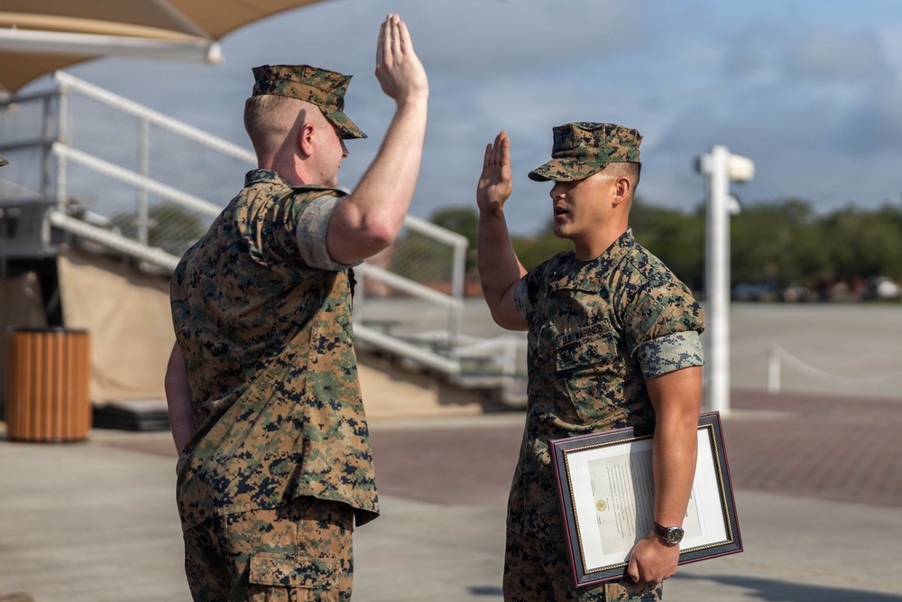 Two U.S. Marines in MARPAT camouflage uniforms are engaged in what appears to be a formal oath or re-enlistment ceremony outdoors on a military installation. The Marine on the right is holding a framed certificate or document while both individuals have their right hands raised. The setting appears to be a parade deck or outdoor ceremonial area.