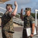 Two U.S. Marines in MARPAT camouflage uniforms are engaged in what appears to be a formal oath or re-enlistment ceremony outdoors on a military installation. The Marine on the right is holding a framed certificate or document while both individuals have their right hands raised. The setting appears to be a parade deck or outdoor ceremonial area.