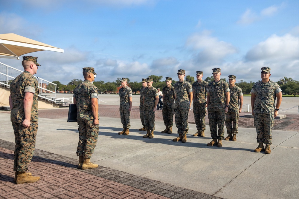A group of military personnel in MARPAT woodland camouflage uniforms are standing in formation on a paved parade deck or drill field. One individual holding a folder or clipboard appears to be addressing or inspecting the assembled group. The setting appears to be a U.S. Marine Corps installation based on the uniforms and environment.