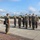 Chief Warrant Officer 2 Joshua J. Krantz’s Promotion Ceremony A group of military personnel in MARPAT woodland camouflage uniforms are standing in formation on a paved parade deck or drill field. One individual holding a folder or clipboard appears to be addressing or inspecting the assembled group. The setting appears to be a U.S. Marine Corps installation based on the uniforms and environment.