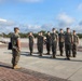 A group of military personnel in MARPAT woodland camouflage uniforms are standing in formation on a paved parade deck or drill field. One individual holding a folder or clipboard appears to be addressing or inspecting the assembled group. The setting appears to be a U.S. Marine Corps installation based on the uniforms and environment.