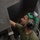 A military aircraft maintainer or technician is inspecting or servicing the underside of a military aircraft. The individual is wearing green hearing protection/earmuffs and an olive green t-shirt, and is looking upward at a panel labeled 'AFT MASTER LATCH (7/32 HEX)'. The close-up shot emphasizes the technical nature of the maintenance work being performed.