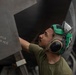 A military aircraft maintainer or technician is inspecting or servicing the underside of a military aircraft. The individual is wearing green hearing protection/earmuffs and an olive green t-shirt, and is looking upward at a panel labeled 'AFT MASTER LATCH (7/32 HEX)'. The close-up shot emphasizes the technical nature of the maintenance work being performed.