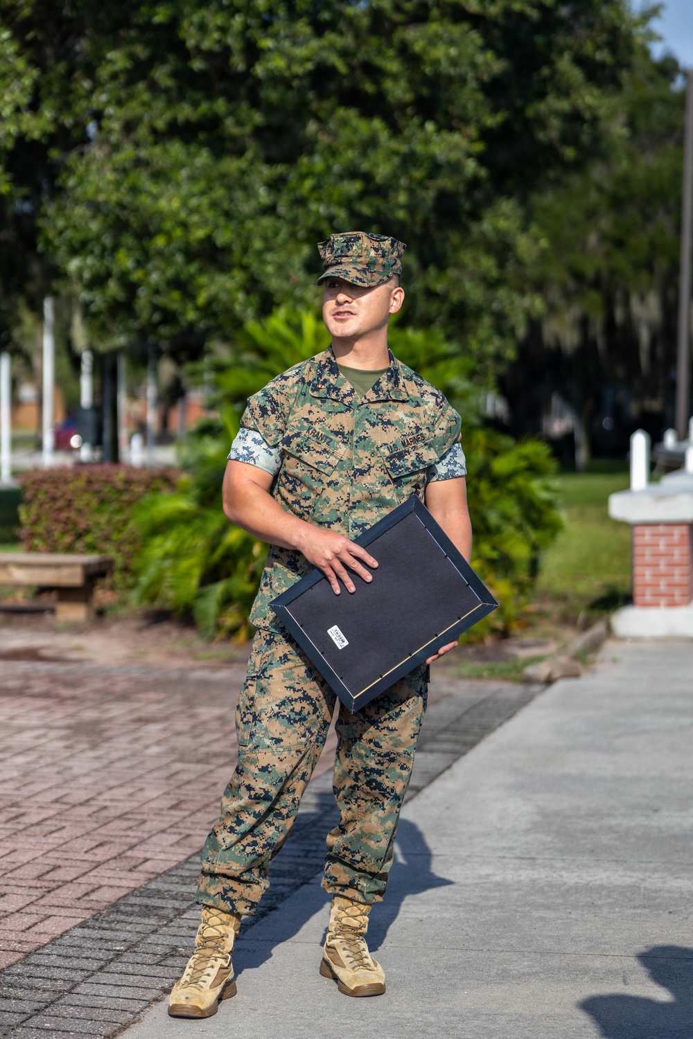 A U.S. Marine in MARPAT woodland camouflage uniform stands outdoors on a paved walkway, holding a dark navy blue presentation or award folder. The Marine's uniform displays a name tape and 'U.S. Marines' chest tape, and they are wearing tan combat boots and a matching camouflage cover (cap). The setting appears to be a military base or installation with brick pathways, manicured landscaping, and trees in the background.