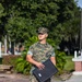 A U.S. Marine in MARPAT woodland camouflage uniform stands outdoors on a paved walkway, holding a dark navy blue presentation or award folder. The Marine's uniform displays a name tape and 'U.S. Marines' chest tape, and they are wearing tan combat boots and a matching camouflage cover (cap). The setting appears to be a military base or installation with brick pathways, manicured landscaping, and trees in the background.