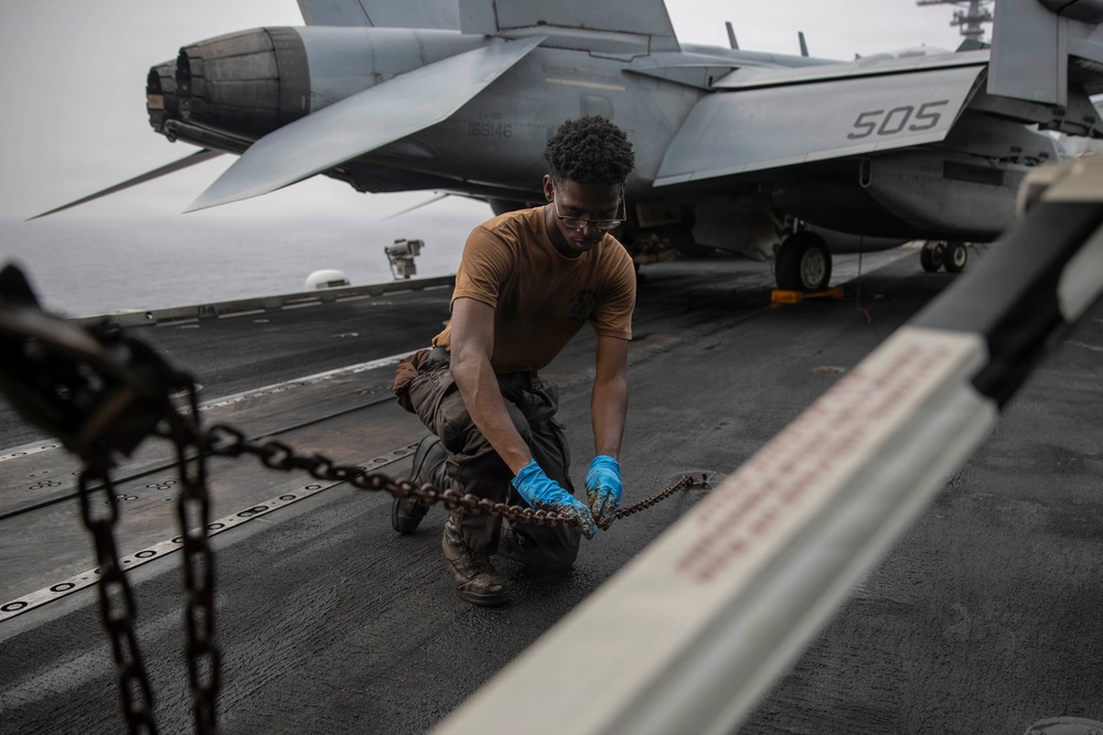 A Navy sailor in a tan shirt and blue gloves is working on the flight deck of an aircraft carrier, handling tie-down chains used to secure aircraft. Behind him is a military jet aircraft marked with the number '505' and bureau number '169148'. The overcast sky and open ocean are visible in the background, indicating the carrier is underway at sea.