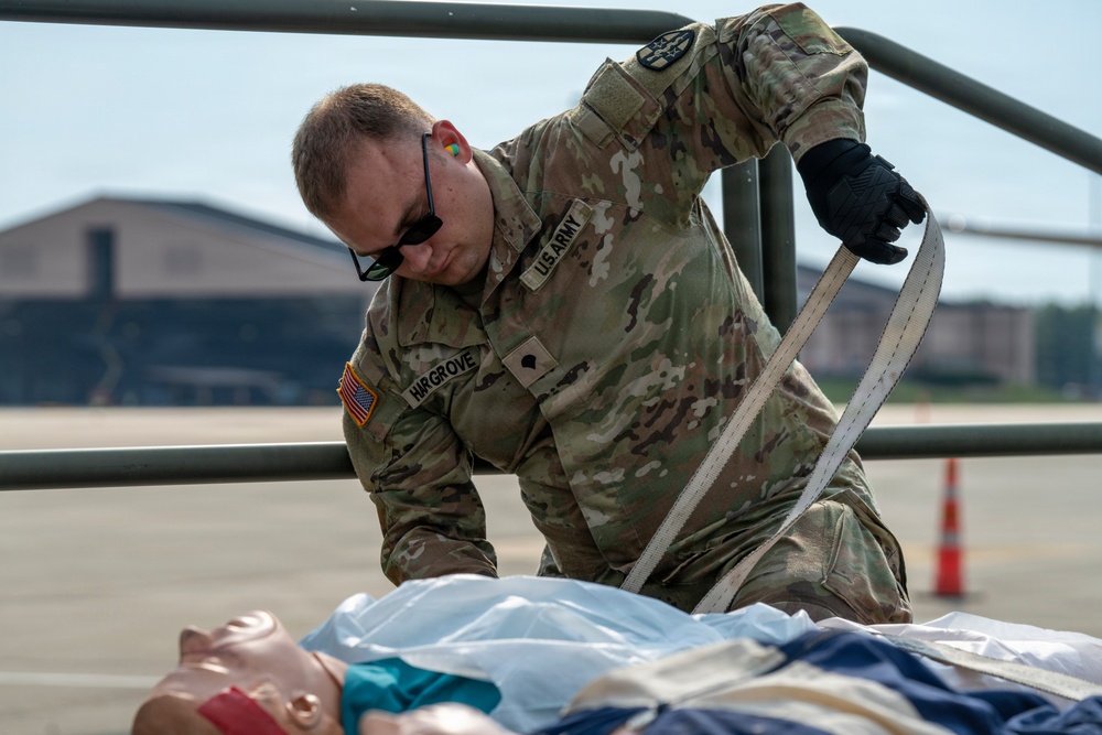 A U.S. Army soldier named Hargrove is conducting a medical training exercise on an airfield tarmac. He is kneeling over a CPR/medical training mannequin and applying what appears to be a stretcher strap or restraint. The setting appears to be a military airfield, with hangars and traffic cones visible in the background.