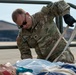 A U.S. Army soldier named Hargrove is conducting a medical training exercise on an airfield tarmac. He is kneeling over a CPR/medical training mannequin and applying what appears to be a stretcher strap or restraint. The setting appears to be a military airfield, with hangars and traffic cones visible in the background.
