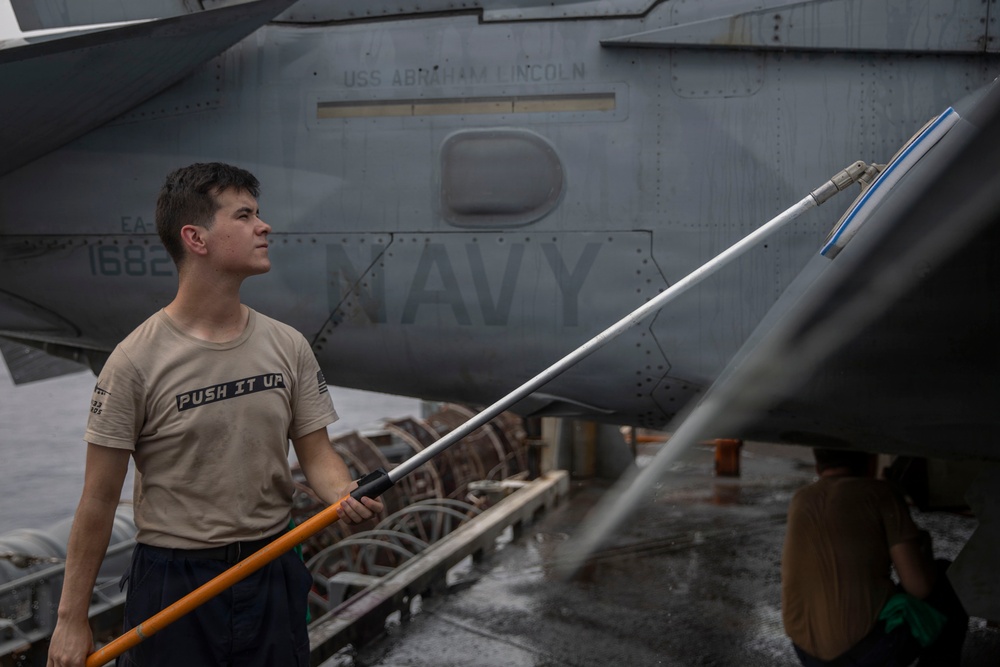A U.S. Navy sailor is cleaning or washing an aircraft aboard the USS Abraham Lincoln aircraft carrier. The sailor is using a long-handled squeegee or brush tool to reach the aircraft's surface. The aircraft visible in the background is marked 'NAVY' and bears the designation 'EA-' with the number '1682', consistent with an EA-18G Growler or similar naval aircraft.