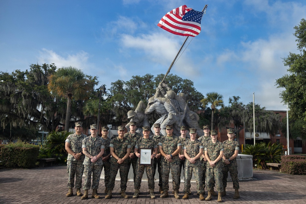 A group of approximately 12 U.S. Marines in woodland MARPAT camouflage uniforms pose for a formal group photograph in front of a replica of the iconic Iwo Jima flag-raising statue. One Marine in the center is holding a framed certificate or award document. The setting appears to be a military installation, likely Marine Corps Recruit Depot (MCRD) Parris Island or a similar base, based on the architecture and landscape.