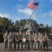 A group of approximately 12 U.S. Marines in woodland MARPAT camouflage uniforms pose for a formal group photograph in front of a replica of the iconic Iwo Jima flag-raising statue. One Marine in the center is holding a framed certificate or award document. The setting appears to be a military installation, likely Marine Corps Recruit Depot (MCRD) Parris Island or a similar base, based on the architecture and landscape.