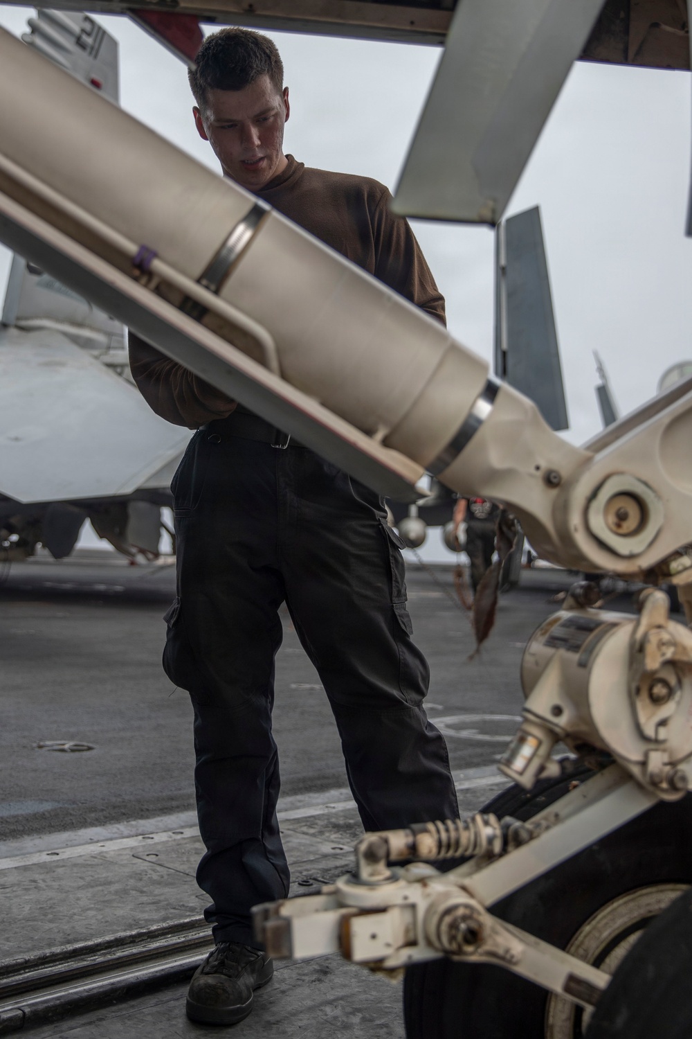 A military personnel member in dark utility clothing is handling what appears to be an air-to-air missile on a weapons loading cart on what appears to be an aircraft carrier flight deck. Fighter jets are visible in the background, consistent with a carrier deck environment. The scene depicts ordnance handling or weapons loading operations.