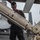 A military personnel member in dark utility clothing is handling what appears to be an air-to-air missile on a weapons loading cart on what appears to be an aircraft carrier flight deck. Fighter jets are visible in the background, consistent with a carrier deck environment. The scene depicts ordnance handling or weapons loading operations.