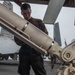 A military personnel member in dark utility clothing is handling what appears to be an air-to-air missile on a weapons loading cart on what appears to be an aircraft carrier flight deck. Fighter jets are visible in the background, consistent with a carrier deck environment. The scene depicts ordnance handling or weapons loading operations.