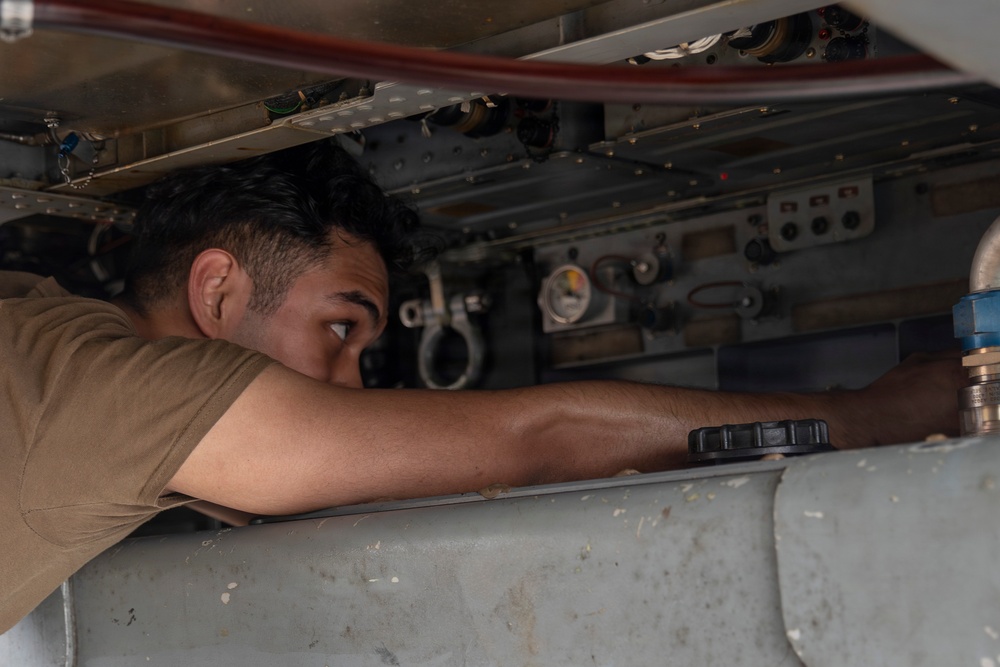 A technician or maintenance worker is inspecting the underside of an aircraft, leaning in closely to examine internal components within an access panel cavity. The individual is wearing a tan/khaki shirt consistent with military or aviation maintenance attire. The image captures a close-up of the worker's face and arm as they peer into the aircraft's mechanical bay.