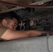 A technician or maintenance worker is inspecting the underside of an aircraft, leaning in closely to examine internal components within an access panel cavity. The individual is wearing a tan/khaki shirt consistent with military or aviation maintenance attire. The image captures a close-up of the worker's face and arm as they peer into the aircraft's mechanical bay.