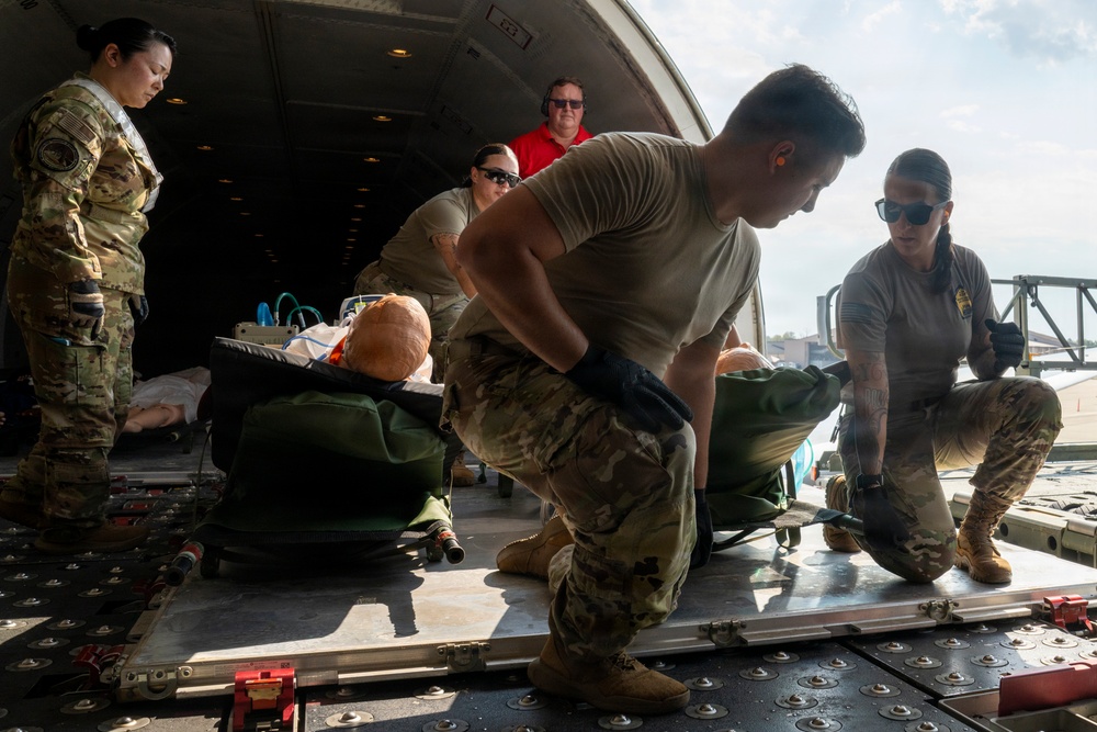 Military personnel in multicam/OCP uniforms are loading or unloading medical litter patients onto or off a military transport aircraft via its rear cargo ramp. The patients appear to be training mannequins, suggesting this is an aeromedical evacuation exercise or training drill. At least five personnel are visible, including one civilian observer in a red shirt wearing hearing protection.
