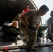Military personnel in multicam/OCP uniforms are loading or unloading medical litter patients onto or off a military transport aircraft via its rear cargo ramp. The patients appear to be training mannequins, suggesting this is an aeromedical evacuation exercise or training drill. At least five personnel are visible, including one civilian observer in a red shirt wearing hearing protection.