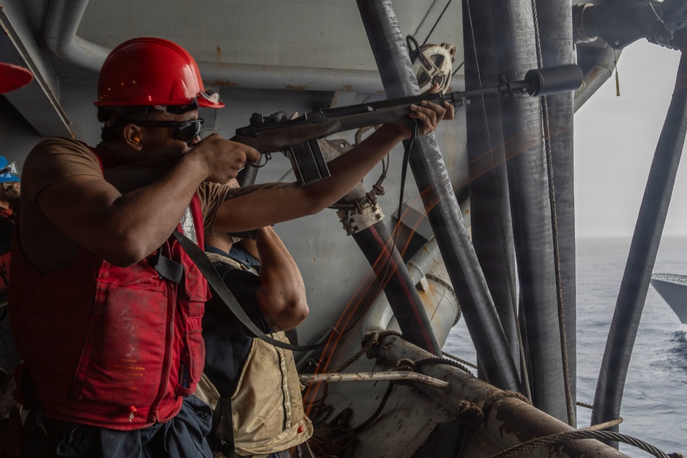 A sailor or military personnel wearing a red hard hat, sunglasses, and a red life vest is aiming a rifle from the deck of a naval vessel. The scene appears to be a live-fire exercise or security drill conducted at sea. Additional crew members are visible in the background, also wearing safety gear.
