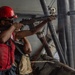A sailor or military personnel wearing a red hard hat, sunglasses, and a red life vest is aiming a rifle from the deck of a naval vessel. The scene appears to be a live-fire exercise or security drill conducted at sea. Additional crew members are visible in the background, also wearing safety gear.