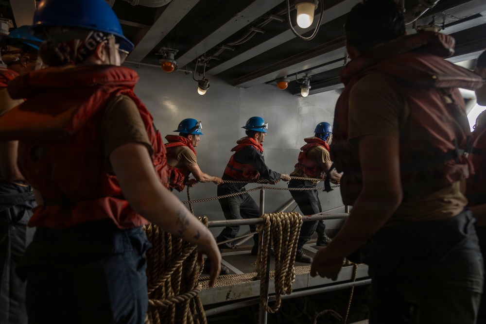 A group of workers or crew members wearing blue hard hats and orange life vests are pulling on thick rope lines aboard what appears to be a naval vessel or large ship. The scene is set in a dimly lit interior deck area with metal structural beams and industrial lighting overhead. The coordinated rope-pulling activity suggests a mooring, line-handling, or seamanship drill or operation.