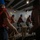 A group of workers or crew members wearing blue hard hats and orange life vests are pulling on thick rope lines aboard what appears to be a naval vessel or large ship. The scene is set in a dimly lit interior deck area with metal structural beams and industrial lighting overhead. The coordinated rope-pulling activity suggests a mooring, line-handling, or seamanship drill or operation.