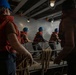 A group of workers or crew members wearing blue hard hats and orange life vests are pulling on thick rope lines aboard what appears to be a naval vessel or large ship. The scene is set in a dimly lit interior deck area with metal structural beams and industrial lighting overhead. The coordinated rope-pulling activity suggests a mooring, line-handling, or seamanship drill or operation.