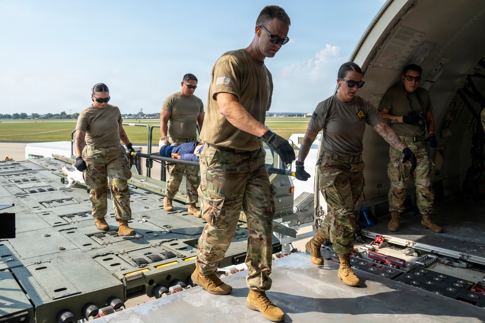 Military personnel in OCP (Operational Camouflage Pattern) uniforms are loading or unloading a litter (stretcher) with a patient/simulated casualty onto or off a military aircraft, likely during a medical evacuation (MEDEVAC) exercise or operation. The scene takes place on an airfield tarmac at the rear cargo ramp of what appears to be a large military transport aircraft. Five service members are actively involved in the patient movement procedure.