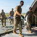 Military personnel in OCP (Operational Camouflage Pattern) uniforms are loading or unloading a litter (stretcher) with a patient/simulated casualty onto or off a military aircraft, likely during a medical evacuation (MEDEVAC) exercise or operation. The scene takes place on an airfield tarmac at the rear cargo ramp of what appears to be a large military transport aircraft. Five service members are actively involved in the patient movement procedure.