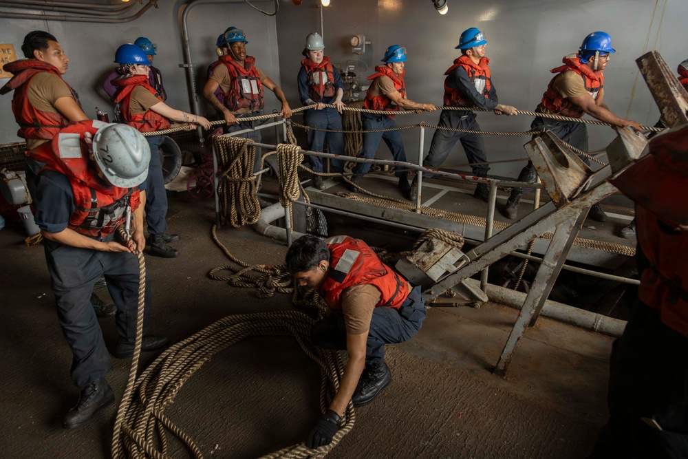 A group of naval personnel are working together on what appears to be a ship's deck or lower compartment, handling thick mooring lines and ropes. The crew members are wearing standard safety gear including blue and white hard hats and orange life vests. The scene depicts a coordinated seamanship evolution, likely a line-handling or mooring drill.