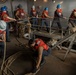 A group of naval personnel are working together on what appears to be a ship's deck or lower compartment, handling thick mooring lines and ropes. The crew members are wearing standard safety gear including blue and white hard hats and orange life vests. The scene depicts a coordinated seamanship evolution, likely a line-handling or mooring drill.