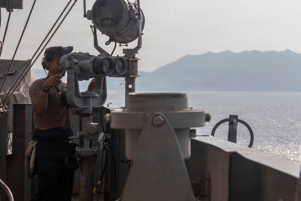 A naval personnel member stands on the bridge wing or deck of a military vessel, using large fixed binoculars to observe the surrounding waters. A mountainous coastline is visible in the background across calm, reflective seas. The scene suggests active maritime surveillance or navigation watch duties.