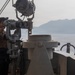 A naval personnel member stands on the bridge wing or deck of a military vessel, using large fixed binoculars to observe the surrounding waters. A mountainous coastline is visible in the background across calm, reflective seas. The scene suggests active maritime surveillance or navigation watch duties.