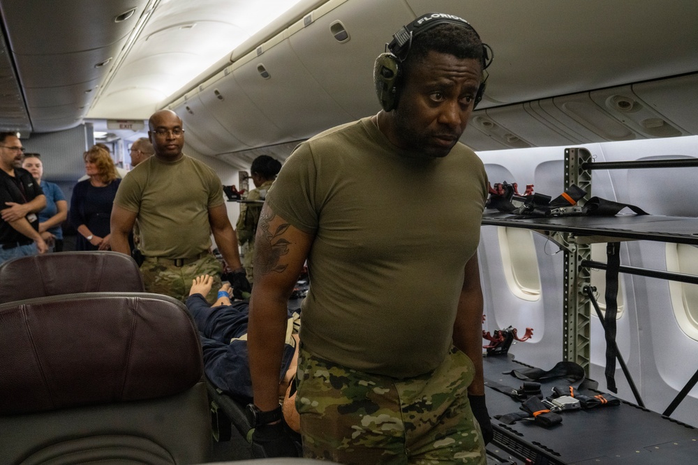 The image shows military personnel in olive drab/multicam uniforms operating inside what appears to be a modified commercial aircraft configured for aeromedical evacuation. A foreground figure wearing hearing protection headsets is positioned near medical litter/stretcher equipment. Several observers in civilian and military attire are visible in the background, suggesting this may be a training exercise or demonstration.