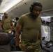 The image shows military personnel in olive drab/multicam uniforms operating inside what appears to be a modified commercial aircraft configured for aeromedical evacuation. A foreground figure wearing hearing protection headsets is positioned near medical litter/stretcher equipment. Several observers in civilian and military attire are visible in the background, suggesting this may be a training exercise or demonstration.