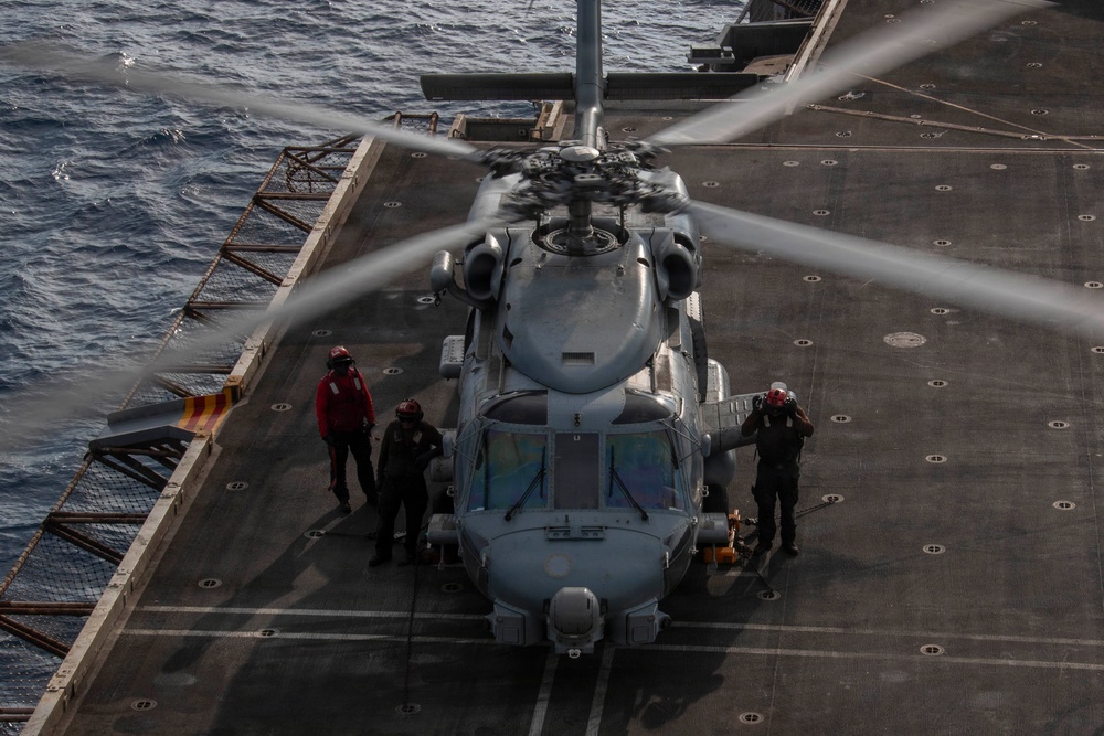 A military helicopter, appearing to be an MH-60 Seahawk or similar naval rotary-wing aircraft, is positioned on the flight deck of a naval vessel at sea. The rotor blades are spinning, indicating the aircraft is either starting up or shutting down. Three deck crew members in flight deck gear are positioned around the aircraft performing ground operations.