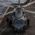 A military helicopter, appearing to be an MH-60 Seahawk or similar naval rotary-wing aircraft, is positioned on the flight deck of a naval vessel at sea. The rotor blades are spinning, indicating the aircraft is either starting up or shutting down. Three deck crew members in flight deck gear are positioned around the aircraft performing ground operations.