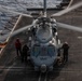 A military helicopter, appearing to be an MH-60 Seahawk or similar naval rotary-wing aircraft, is positioned on the flight deck of a naval vessel at sea. The rotor blades are spinning, indicating the aircraft is either starting up or shutting down. Three deck crew members in flight deck gear are positioned around the aircraft performing ground operations.