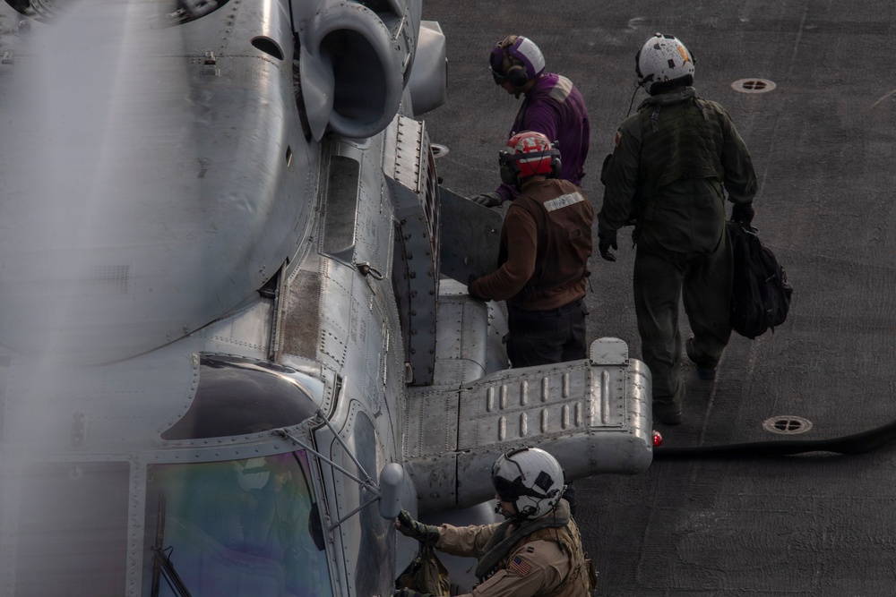 Military personnel are conducting pre-flight or post-flight operations on a naval flight deck alongside a military helicopter, likely a CH-53 or similar heavy-lift rotary-wing aircraft. At least four individuals are visible, wearing different colored helmets and flight gear indicating various crew roles. The scene appears to take place on the deck of a naval vessel.